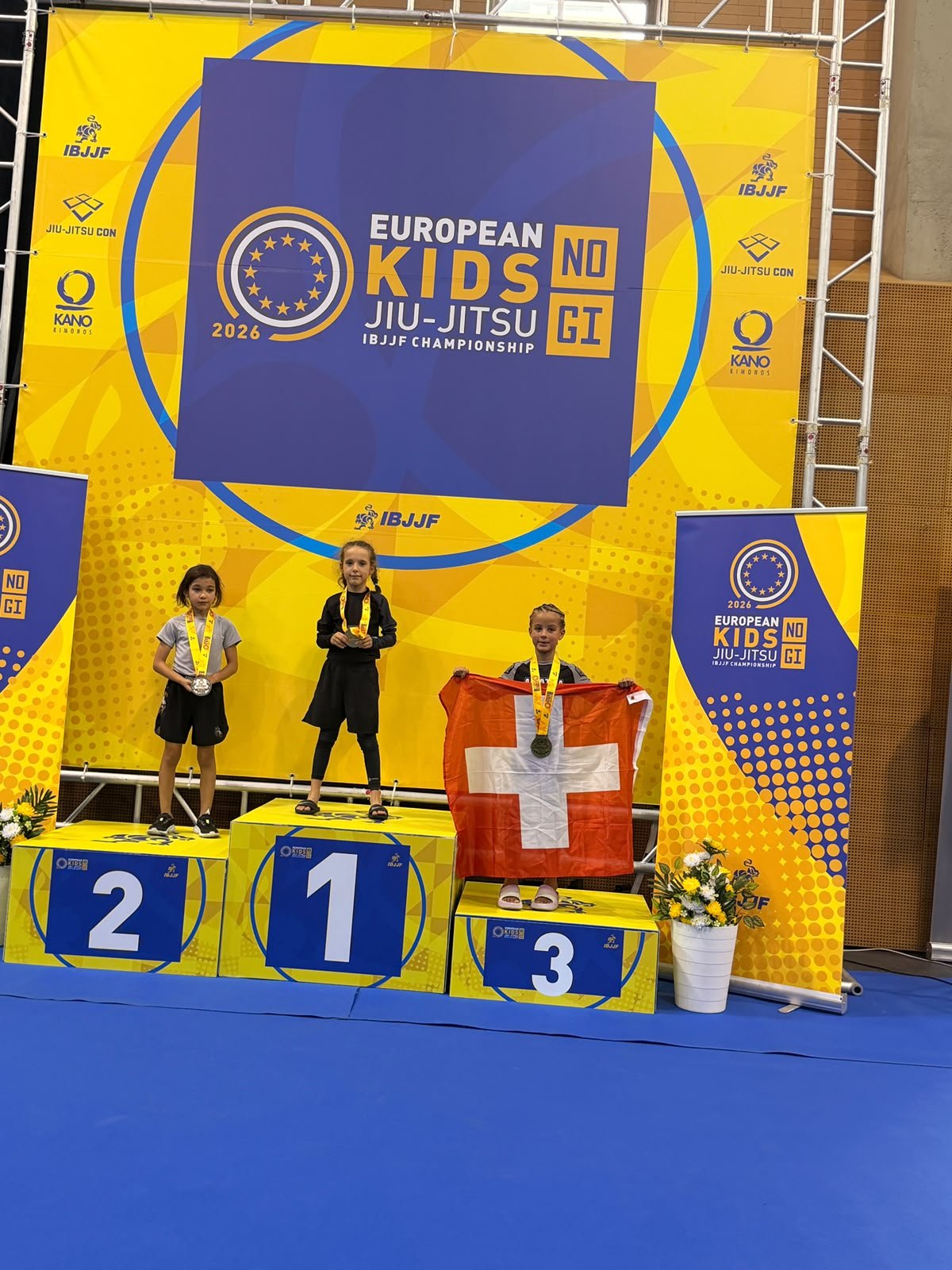Aleyna auf dem dritten Podestplatz mit der Schweizer Fahne an der European Kids No-Gi IBJJF Championship, Lissabon, Oktober 2026.